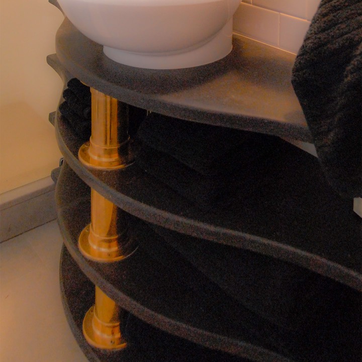 three black soapstone shelves curve along a bathroom wall, supported at their greatest extension by thick copper pipes